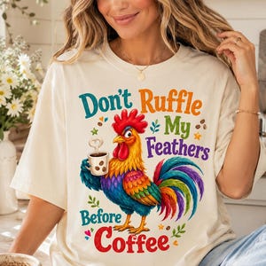 Pode incluir: Camiseta creme com um galo colorido segurando uma xícara de café. O galo tem uma crista vermelha, um bico amarelo e penas vibrantes azuis, verdes e roxas. O texto diz "Don't Ruffle My Feathers Before Coffee."