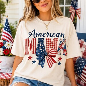 Pode incluir: Camiseta creme com o texto "American MAMA" em designs patrióticos vermelhos, brancos e azuis. O design inclui estrelas, listras e um laço. A camisa tem um ajuste casual e descontraído.