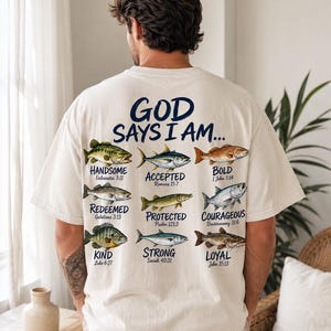 Pode incluir: Camiseta creme com a frase "GOD SAYS I AM..." e várias ilustrações de peixes. Cada peixe é combinado com uma afirmação positiva e um versículo bíblico correspondente. O design da camiseta inclui as palavras "Handsome", "Accepted", "Bold", "Redeemed", "Protected", "Courageous", "Kind", "Strong" e "Loyal".