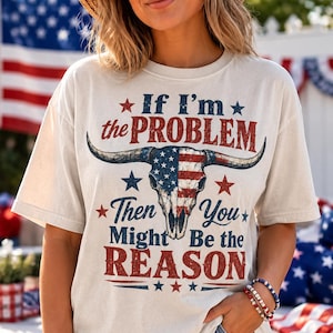 May include: Cream-colored t-shirt with the text "If I'm the PROBLEM Then You Might Be the REASON" in red and blue. The graphic features a bull skull with an American flag design. The shirt is short-sleeved.