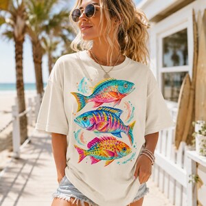 May include: An off-white short-sleeve t-shirt featuring a graphic print of three colorful fish stacked vertically. The fish are illustrated in vibrant shades of pink, blue, yellow, and orange with watercolor-style splashes surrounding them.
