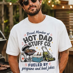 May include: A white short-sleeve t-shirt featuring a graphic print with the text Proud Member of HOT DAD STUFF CO. and FUELED BY propane and bad jokes. The design includes illustrations of a charcoal grill, a propane tank, a cooler, and a lawnmower.