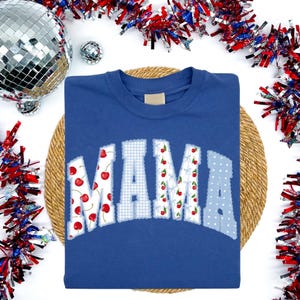 May include: A blue t-shirt with the word "MAMA" appliqued across the front. The letters are in a mix of patterns, including cherries, gingham, and polka dots. The shirt is displayed on a woven mat with festive decorations.