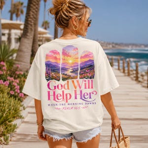 Pode incluir: Uma t-shirt de manga curta em tom off-white com um gráfico nas costas de um nascer do sol colorido sobre montanhas e flores. O texto diz God Will Help Her WHEN THE MORNING DAWNS PSALM 46:5 em letras com gradiente rosa, laranja e roxo.