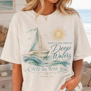Puede incluir: Una camiseta de manga corta color hueso con un gráfico en acuarela de un velero sobre olas azules y un sol amarillo. El texto dice: WHEN YOU PASS THROUGH Deep Waters I Will Be With You ISAIAH 43:2, acompañado por un pequeño icono de ancla.