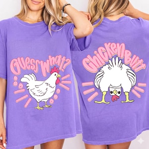 Może przedstawiać: Fioletowa koszulka z zabawnym motywem kurczaka. Przód przedstawia napis "Guess What?" i ilustrację kurczaka. Tył pokazuje tyłek kurczaka z napisem "Chicken Butt". Tekst i wzór są w kolorze różowym i żółtym.