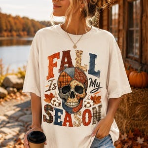 Peut inclure: Un t-shirt à manches courtes blanc cassé avec un motif de crâne au design patchwork. Le texte "FALL IS MY SEASON" est imprimé en grandes lettres capitales multicolores autour du crâne, agrémenté d'illustrations de feuilles d'automne.