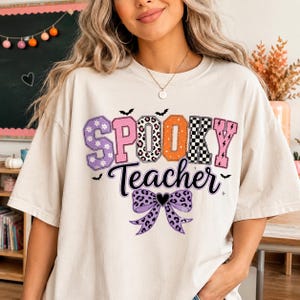 Könnte beinhalten: Ein cremefarbenes T-Shirt mit dem Schriftzug 'SPOOKY Teacher' in einer gemusterten Schriftart. Die Buchstaben zeigen Blumen-, Leoparden-, orangefarbene, karierte und Blitz-Muster. Unter dem Text befindet sich eine violette Leoparden-Schleife.