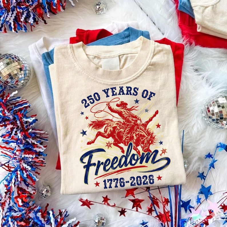 May include: A stack of t-shirts in red, white, and blue. The top shirt is cream-colored with a graphic of a cowboy on a bucking bronco, the text "250 Years of Freedom 1776-2026". Decorative elements include stars and disco balls.