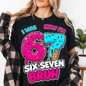 May include: Black t-shirt with a colorful graphic that says "I WAS NOW I'M 67 SIX-SEVEN BRUH". The number 6 is designed as a pink donut with sprinkles, and the number 7 is designed as blue ice cream.