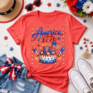 May include: Red t-shirt with a patriotic design featuring the word "America" and the year "1776". The graphic includes a cake with candles, fireworks, and an American flag. The image also includes denim shorts, sunglasses, and a straw hat.