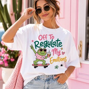 Op de afbeelding: Een wit t-shirt met een afbeelding van een groene kikker met een hartvormige zonnebril, een geruit overhemd en op een roze skateboard. De kikker houdt een drankje vast, met de tekst: Off To Regulate My Emotions, vergezeld door sterren en een hart.