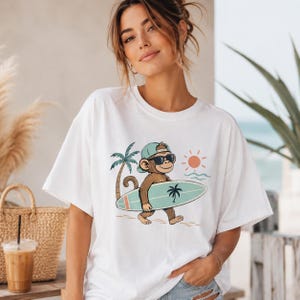 Könnte beinhalten: Ein weißes Kurzarm-T-Shirt mit einer grafischen Illustration eines braunen Affen, der eine hellblaue Baseballkappe und eine schwarze Sonnenbrille trägt. Der Affe trägt ein hellblaues Surfbrett mit Palmenmotiv neben einer Palme und Sonne.