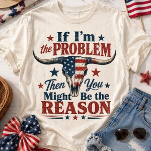 May include: Cream-colored t-shirt with the text "If I'm the PROBLEM Then You Might Be the REASON" and a bull skull design in red, white, and blue. The shirt is paired with denim shorts, sunglasses, and patriotic decorations.