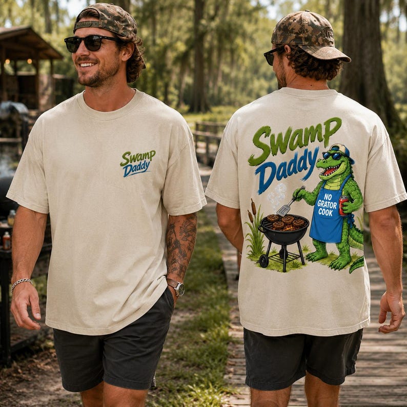 May include: Beige t-shirt with "Swamp Daddy" logo on the front and back. The back features a cartoon alligator grilling burgers with the text "No Gator Cook". The shirt is worn with dark shorts and a camouflage cap.