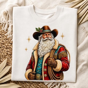 Puede incluir: Una camiseta blanca con el gráfico de una figura barbuda que lleva un sombrero de vaquero marrón con acebo, un abrigo rojo con ribete de piel, un chaleco azul y guantes marrones. La figura sostiene una cuerda y hay estrellas doradas.
