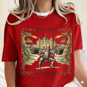 May include: Red t-shirt featuring a medieval knight lifting a barbell in a squat position. The design includes a castle, dragons, and the text "Dost thou even hoist?" in a decorative frame.