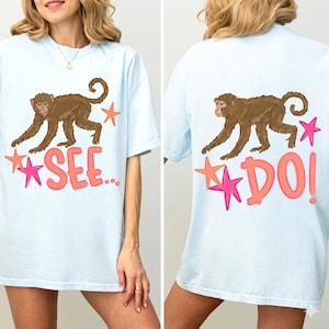 Op de afbeelding: Lichtblauw T-shirt met een bruine apenillustratie en de tekst "SEE..." op de voorkant. De achterkant van het shirt heeft dezelfde apenillustratie en de tekst "DO!". Roze en oranje steraccenten zijn inbegrepen.