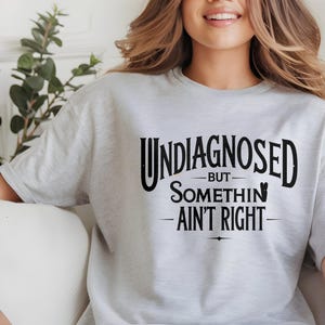Op de afbeelding: Een grijs T-shirt met zwarte tekst die "Undiagnosed - But somethin' ain't right" luidt.