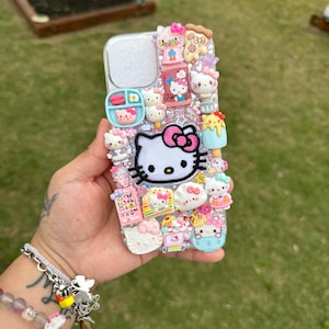 Może przedstawiać: Srebrne etui na telefon zdobione licznymi zawieszkami 3D, w tym dużą postacią Hello Kitty z r&oacute;żową kokardą. Inne zawieszki przedstawiają jedzenie i figurki Hello Kitty. Etui ma przezroczysty otw&oacute;r na aparat i jest ozdobione r&oacute;żnymi uroczymi ozdobami.