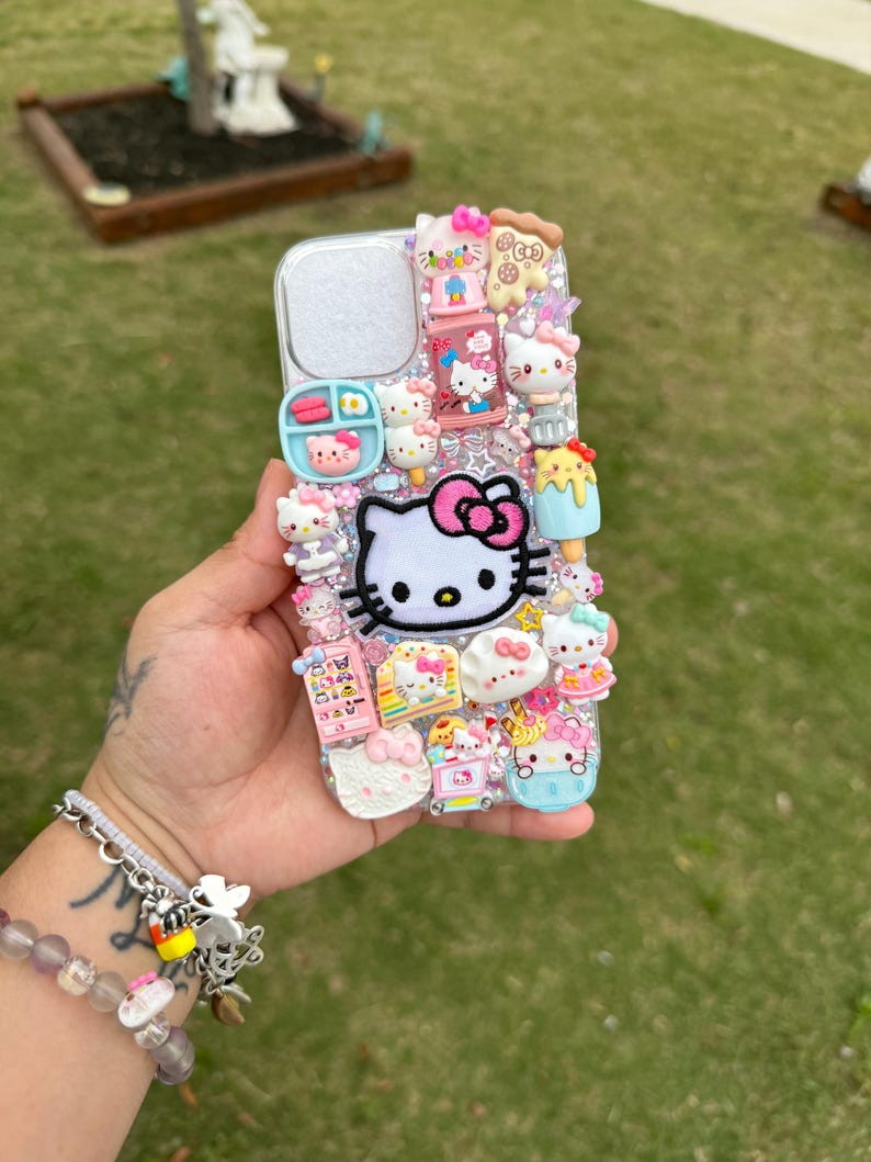 Może przedstawiać: Srebrne etui na telefon zdobione licznymi zawieszkami 3D, w tym dużą postacią Hello Kitty z r&oacute;żową kokardą. Inne zawieszki przedstawiają jedzenie i figurki Hello Kitty. Etui ma przezroczysty otw&oacute;r na aparat i jest ozdobione r&oacute;żnymi uroczymi ozdobami.