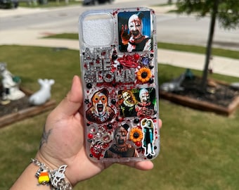 May include: A clear phone case decorated with Art the Clown imagery, featuring silver glitter text that reads ART THE CLOWN, small orange sunflower charms, red and black beads, and various horror-themed character stickers.