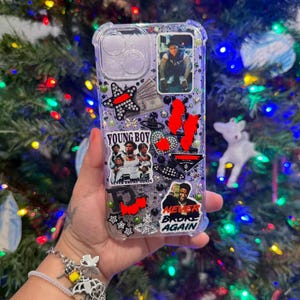 Può includere: Custodia trasparente per telefono con glitter e abbellimenti decorativi. Presenta immagini di una persona, una stella, denaro e i testi "YOUNG BOY" e "NEVER BROKE AGAIN". La custodia è tenuta davanti a un albero di Natale.