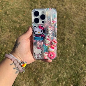 Może przedstawiać: Przezroczyste etui na telefon ozdobione figurkami Hello Kitty, kokardkami i innymi ozdobami. Etui posiada r&oacute;żnorodne r&oacute;żowe, niebieskie i białe akcenty, w tym r&oacute;żowy pączek i gitarę. Etui jest trzymane w dłoni.