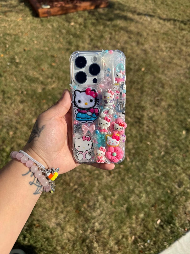 Może przedstawiać: Przezroczyste etui na telefon ozdobione figurkami Hello Kitty, kokardkami i innymi ozdobami. Etui posiada r&oacute;żnorodne r&oacute;żowe, niebieskie i białe akcenty, w tym r&oacute;żowy pączek i gitarę. Etui jest trzymane w dłoni.