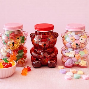 Puede incluir: Tres tarros de caramelos con forma de oso llenos de golosinas coloridas. Un tarro contiene ositos de goma, otro está lleno de caramelos rojos y el tercero contiene caramelos en forma de corazón. Los tarros tienen tapas rosas o rojas.