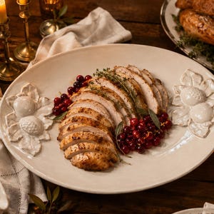 May include: An oval-shaped white ceramic serving platter featuring raised relief details of acorns and oak leaves on the handles. The platter is filled with slices of roasted poultry, garnished with fresh green herbs and bright red cranberries.