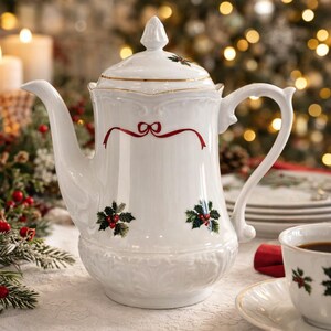 May include: A white porcelain coffee pot with a lid, red bow detail, and holly berry accents. A matching teacup and saucer are on the table, along with a stack of plates. The scene is set for a festive holiday gathering.