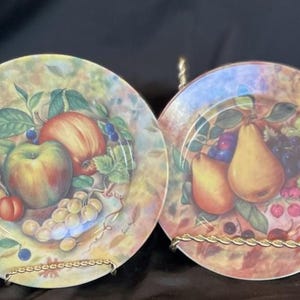 May include: Four decorative plates with fruit designs, including plums, apples, pears, and peaches. Each plate is displayed on a gold-colored metal stand. The plates have a colorful, detailed design.