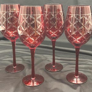 May include: Four ruby red wine glasses with diamond-cut patterns. The glasses have long stems and round bases. The word "CELLINI" is visible on one glass. The glasses are arranged on a dark surface.