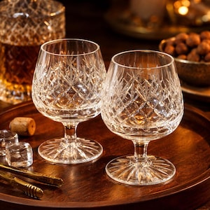 Puede incluir: Dos copas de brandy de cristal tallado con patrones de diamantes sobre una bandeja de madera. Una jarra, cubitos de hielo y una vela encendida en el fondo crean un ambiente cálido. Las copas están diseñadas para disfrutar de licores.