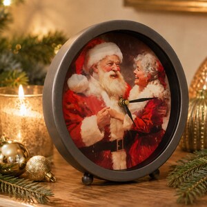 May include: A round analog clock with a dark metal frame and small feet. The clock face features a festive illustration of Santa Claus and Mrs. Claus in red outfits, smiling at each other. Black clock hands are positioned in the center of the image.