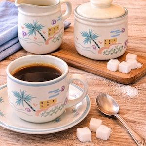 Peut inclure: Un ensemble tasse et soucoupe à café, pot à crème et sucrier avec un motif tropical. Les pièces en céramique blanche présentent un palmier, des vagues et un petit bâtiment aux couleurs pastel. Une cuillère et des cubes de sucre sont également présents.