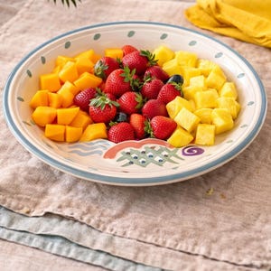 May include: A decorative ceramic plate filled with fresh fruit. The plate is divided into sections of diced mango, strawberries, and pineapple. The plate has a blue and white rim and decorative painted elements.