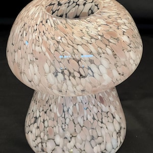 May include: A decorative glass vase with a mushroom shape. The vase is a light pink color with white and black speckles. The top of the vase has a wide opening. The vase is a unique piece of home decor.
