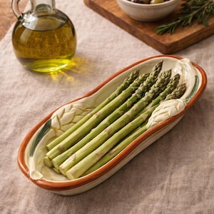 May include: A ceramic serving dish filled with fresh green asparagus spears. The dish has a cream-colored interior with a brown rim and leaf-shaped accents. A glass bottle of olive oil and a bowl of olives are also present.