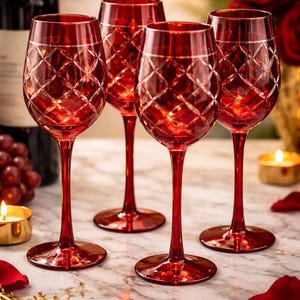 Puede incluir: Cuatro copas de vino rojas con detalles de rombos. Las copas tienen tallos largos y están sobre una superficie de mármol. El fondo incluye una botella de vino, uvas, velas y pétalos de rosa.
