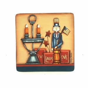 May include: A painted wooden square with a patriotic theme. A figurine of a person wearing a blue jacket, red hat, and holding a flag stands on a wooden base. The base is surrounded by three red boxes with the words "Live", "Laugh", and "Love" written on them. A silver candlestick with two lit candles is in the background.