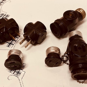 May include: A collection of six vintage black electrical adapters with various configurations, including a three-prong adapter, a two-prong adapter, and a light bulb socket adapter.