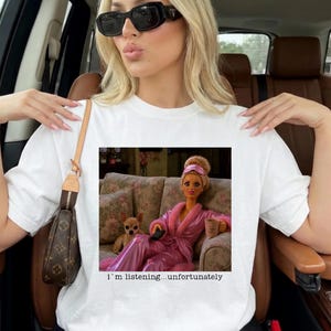 Op de afbeelding: Een wit T-shirt met korte mouwen en een grafische print van een pop in een roze badjas en een kleine hond op een bank met bloemenpatroon. De pop houdt een mok vast en draagt een slaapmasker. Onder de afbeelding staat: i'm listening...unfortunately.