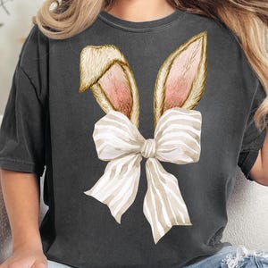 May include: A dark gray t-shirt with a watercolor illustration of bunny ears and a white and brown striped bow.