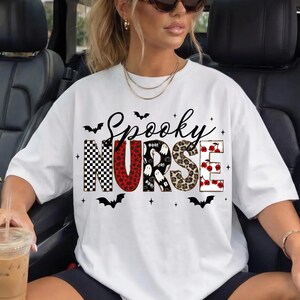 May include: A white short-sleeve t-shirt featuring the text Spooky Nurse in a large, multi-patterned font. The letters include black and white checkerboard, red leopard print, white ghosts on black, brown leopard print, and red cherries on white.