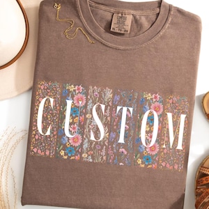Puede incluir: Una camiseta marrón con estampado floral y la palabra "CUSTOM" en letras blancas. La camiseta está hecha por Comfort Colors y es talla XL.