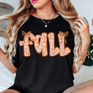 May include: Black t-shirt with the word "FALL" in orange, floral-patterned letters. The letters are outlined in orange and have orange bows at the top. The floral pattern includes pink and orange flowers.