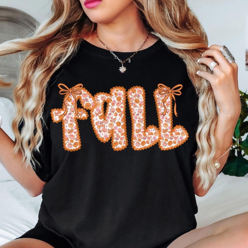 May include: Black t-shirt with the word "FALL" in orange, floral-patterned letters. The letters are outlined in orange and have orange bows at the top. The floral pattern includes pink and orange flowers.