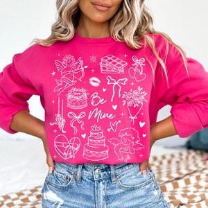 May include: A bright pink sweatshirt with white graphic illustrations of hearts, bows, cakes, and cherubs. The words "Be Mine" are written in a script font. The sweatshirt is a crew neck style.